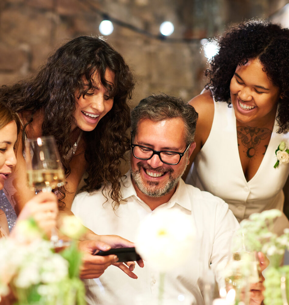 Happy young brunette bride showing her parents and friend new photos in smartphone while bending over cheerful mature man
